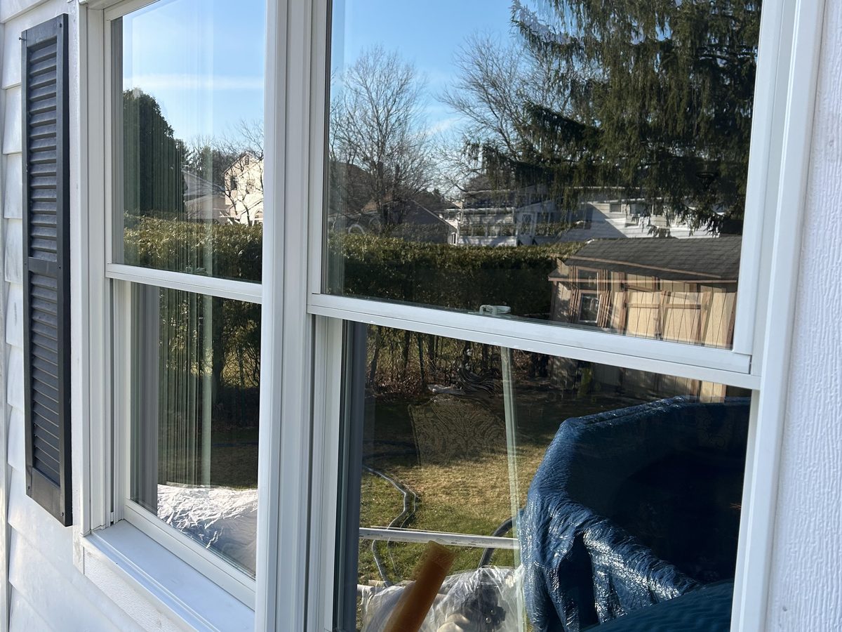 A clean double-hung window on a white-sided house, reflecting blue sky and pine trees
